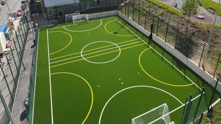 A multi-use games area with a green astro-turf pitch with white and yellow semi-circles on it and a large green fence around its edge.