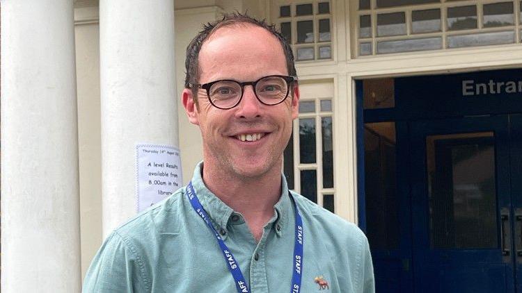 A white middle aged man with black rimmed glasses and wearing a green shirt and a blue staff lanyard smiles at the camera outside Ashcombe School. 