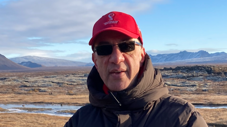 Keith Georgiou standing in front of a beach. They are wearing a red cap, sunglasses and a brown coat 