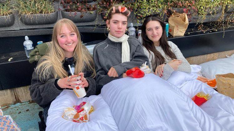 Lucy (left) has long blonde hair, Evie has light bow hair which is in rolles, Erin has long brown hair. All thee are wearing grey jumpers, they are sitting on the floor under a duvet, eating McDonald's.