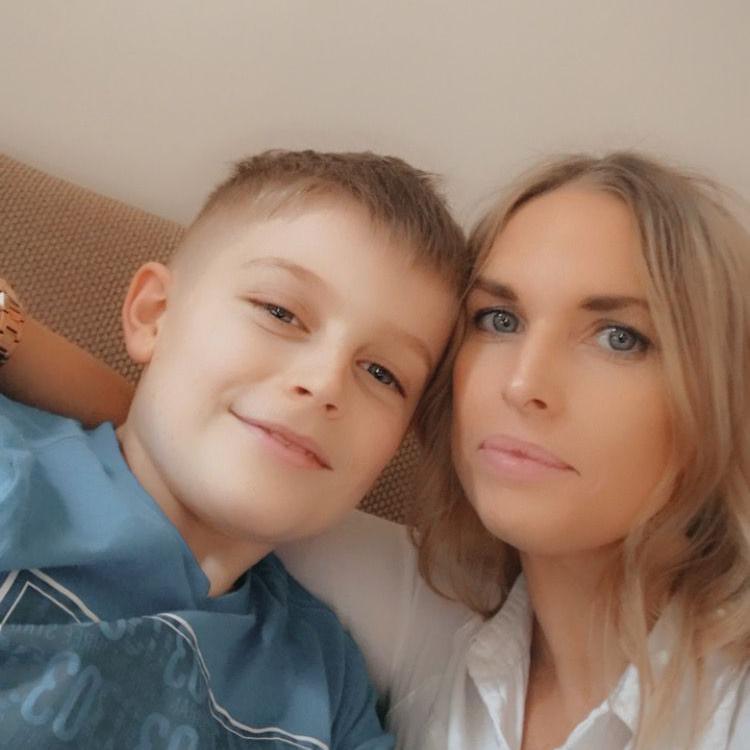 A young boy with blond hair smiles at the camera. His head is leaned in towards his mother, a blonde woman with blue eyes who looks at the camera.