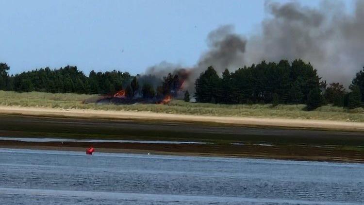 Black smoke rising from fire on marram grass in Wells-next-the-Sea, Norfolk, with water in front