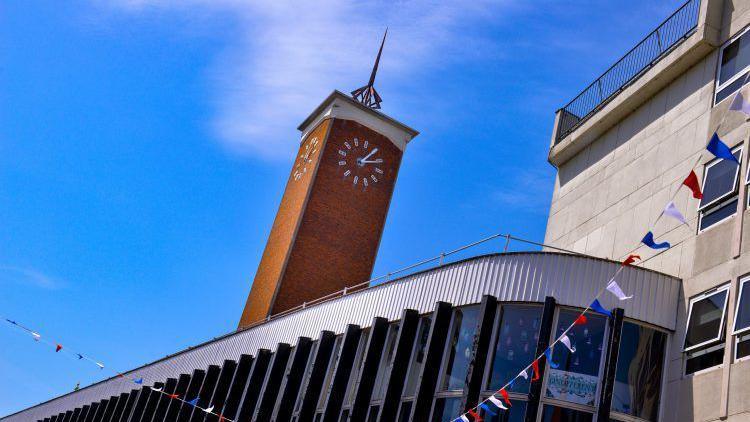 The top of a 1960s postmodern building, with red brick clock tower and spire. There is red, whiteand blue bunting attached to the structure and a blue sky as its backdrop.