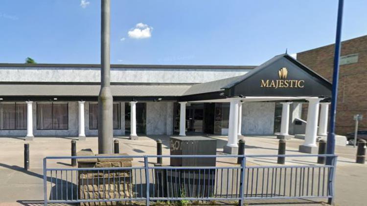 A long white one story marble covered building with several support pillars, behind street furniture including street lights and a grey metal railings fence.