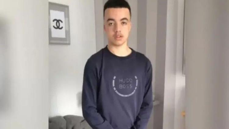 A teenage boy with short, dark hair. He is stood up and is wearing a navy, long-sleeved top with Hugo Boss written on the front. 