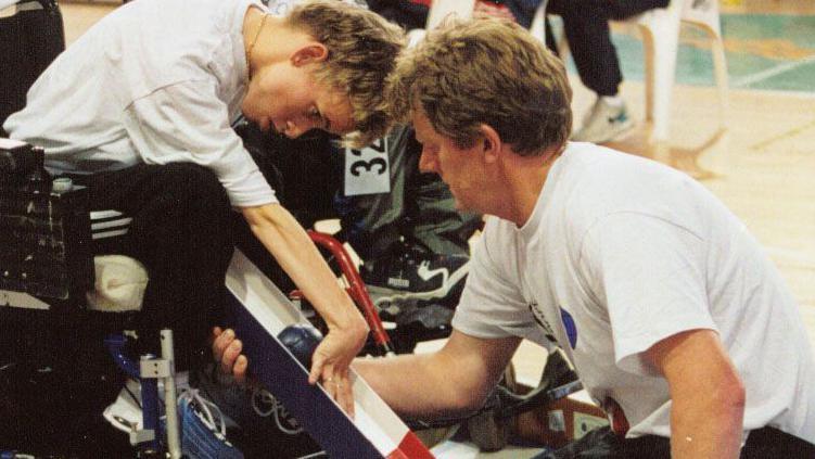 A woman in a wheelchair roles a navy ball down a colourful tube supported by a man kneeling in a white t shirt