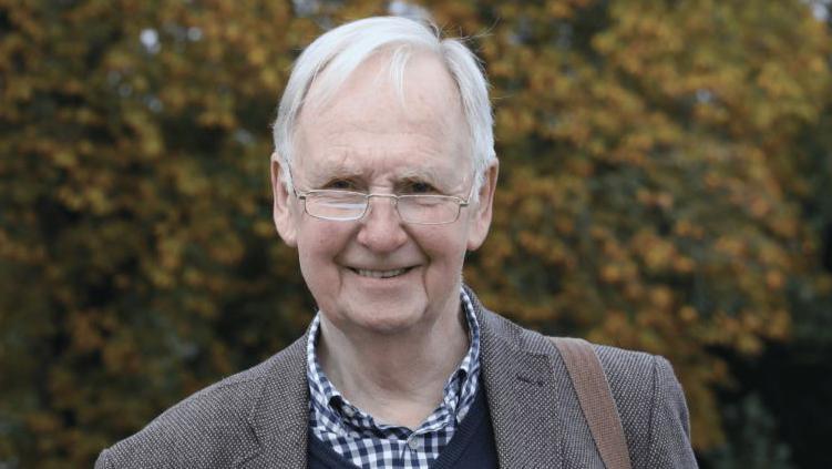 John Smith in a blazer with a blue chequered shirt. He is wearing eye glasses and has grey hair.