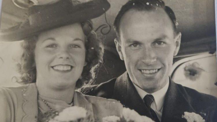 Man and woman are sitting in a car and both are smiling. Man wears a suit with tie. His wife has a hat, carries flowers and is also in a light-coloured suit. The image is a black and white photo.