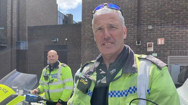 In the foreground is David - he's wearing a bright yellow jacket, with a stripe across the middle in blue and white checks. He's got a pair of blue sunglasses on his head and is wearing a black jacket underneath. Behind him, to his right, is Andy, who's wearing the same jacket. They're both sat on police motorbikes, and behind them is a dark brick building. 