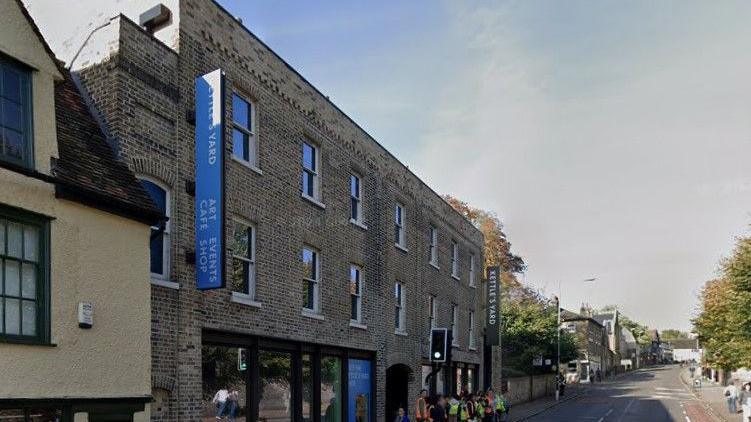 View of the front of Kettle's Yard gallery in Cambridge. It is a brick building with banners and signs on it showing its name. It is on a busy city street and there are a number of school children outside with a teacher, waiting at a pedestrian crossing