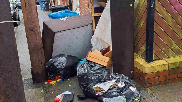 Three black bin bags dumped on a street, one with Lego spilling out of it. Two large brown wooden bedframes are dumped next to the bags, with one standing upright and one on its side