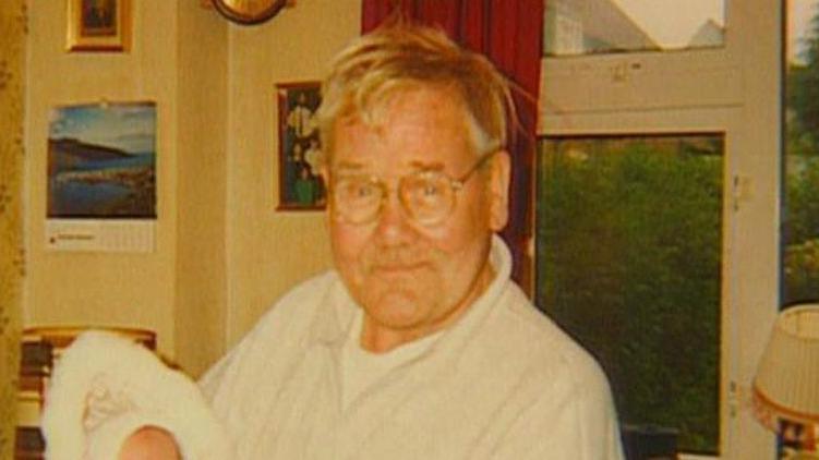 A man aged approximately in his late 70s hold a baby that is just out of sight in his arms.