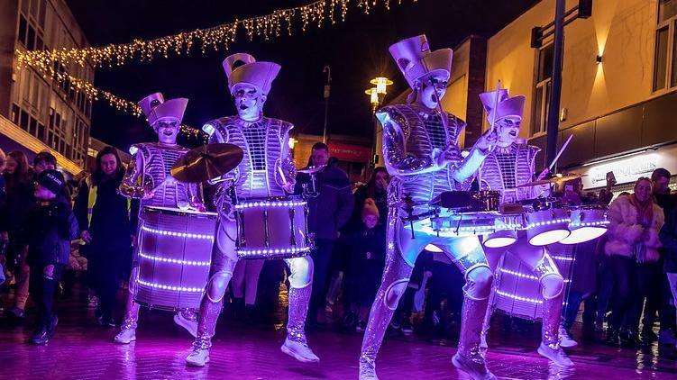 Four people in white costumes and covered in lights perform outside. They are surrounded by people watching on. 