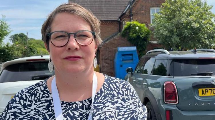 Dawn Thomas is wearing a blue top with white leaf patterns. She has light brown hair, of medium length, and she is wearing glasses. She is pictured near to the school building, with cars parked nearby.