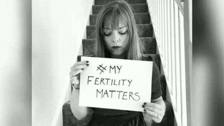 A black and white photo of Sarah Curtiss sitting on stairs in a home and holding a hand-written sign reading "#My Fertility Matters".