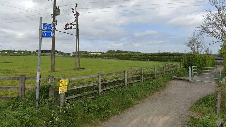 A cycle track bordered by a fence, then hedgerows, with a blue sign pointing towards a five-bar gate beyond which the track continues. There is a green field to the left.