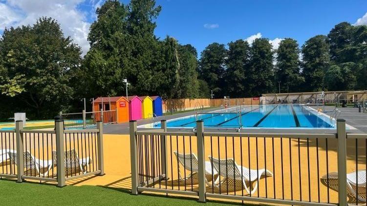 An outside pool with lanes. In the foreground there are white deck chairs. To the left of the pool there are four beach huts, painted orange, pink, yellow and blue.