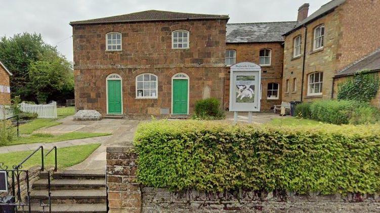 Two-storey stone-built chapel with two green doors on either side of a arched window on the ground floor and two arched windows above, with small square pains of glass separated by white frames. There is a grassed area in front, criss-crossed by paths and another stone building to the right. There is a hedge in the foreground and some stone steps with railings.