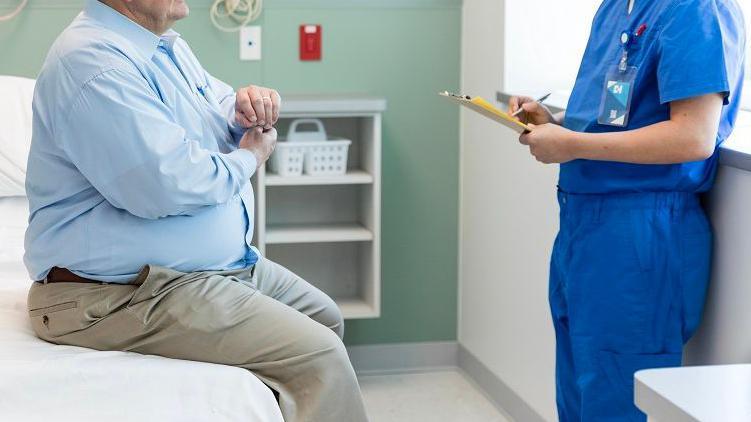 An overweight man sits on a bed in a clinical setting, in consultation with a medic wearing blue overalls. Their faces are cropped out of the picture