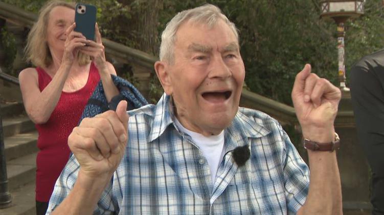 George has his hands in the air and is cheering. He is wearing a check shirtand has grey hair.