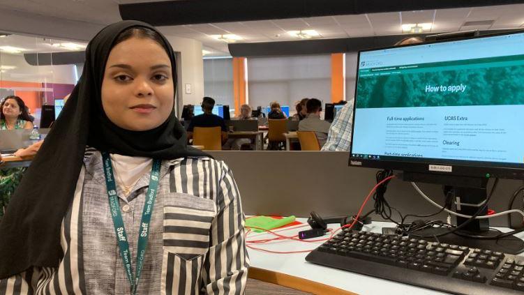 A woman sits at a desk in front of a computer in a call centre, she is wearing a black headscarf.