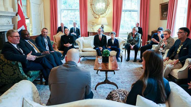 Keir Starmer meets families of hostages held in Gaza - He is seen sitting next to David Lammy on a sofa, with 13 people sat on sofas surrounding them in a discussion. A Union Flag is visible in the room, with peachy red curtains on the windows and what looks like a mahogany table in the middle of the room