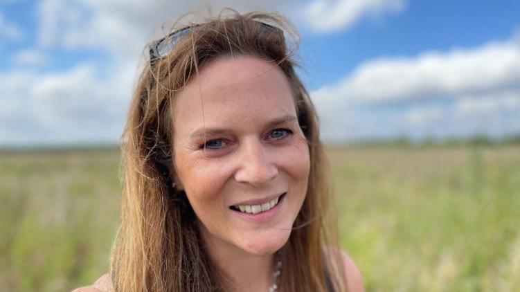 Hannah Dale with long brown hair smiling and looking into the camera with the farm in the background 