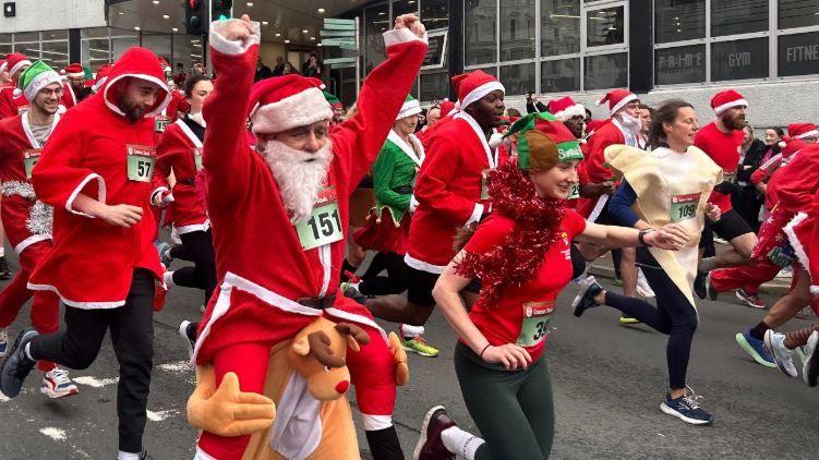 Lots of people in red Santa suits and trainers run along a road. A man with a fake white beard and a Santa hat lifts his hands up in the air.