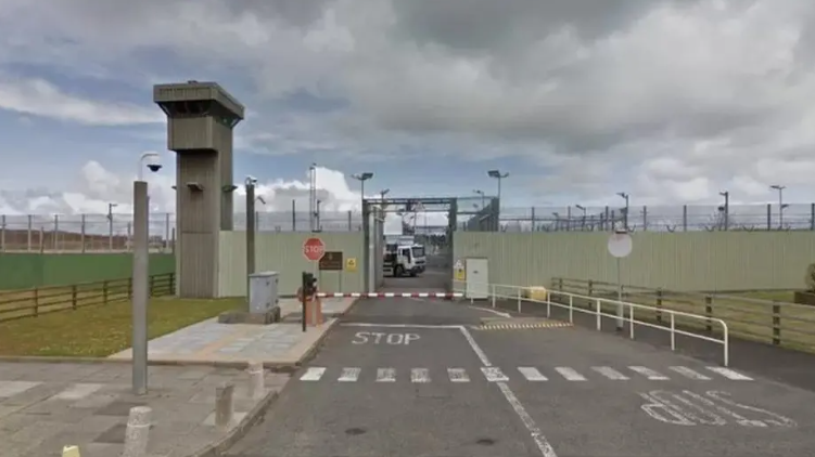 Shows an open prison gate with a barrier to the front and a security tower to the left, with a white prison van on the inside.