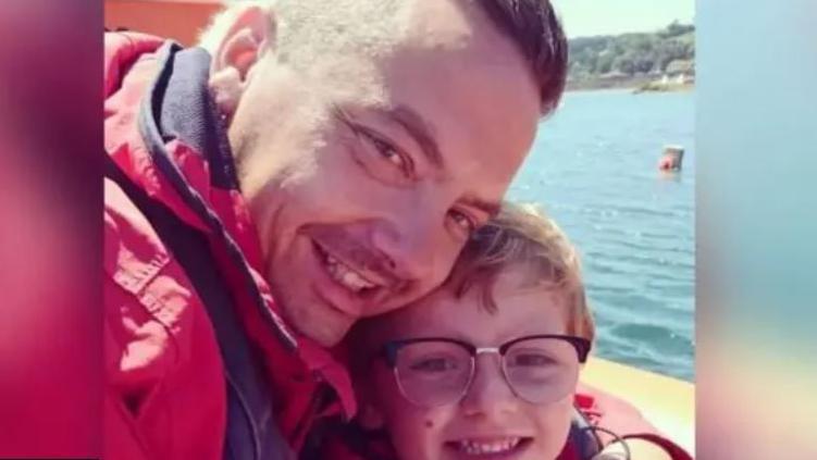 Dean and Charlie Lowe posing together in a selfie with both smiling and wearing red coats. They are on a boat.