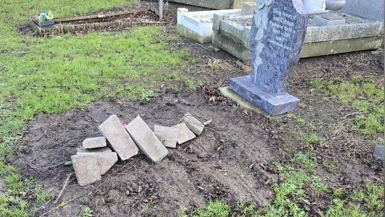 Four headstones can be seen on a patch of grass. The nearest grave site is muddy and has tyre tracks over it. A mound of bricks has been put on top of the grave.