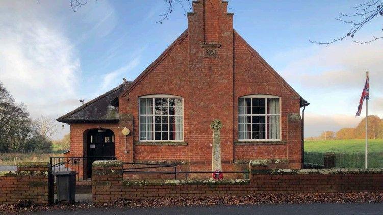 Scaleby Hall, a small red brick building, with the war memorial outside it.