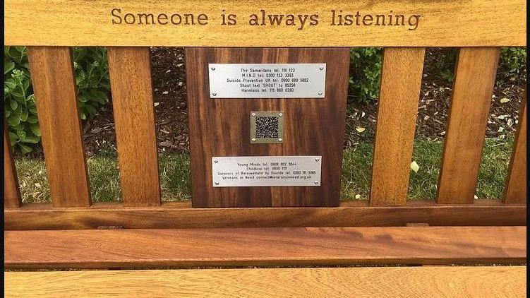 A wooden bench. Three small metal plates are inscribed with numbers and a QR code carrying details of mental health support groups.