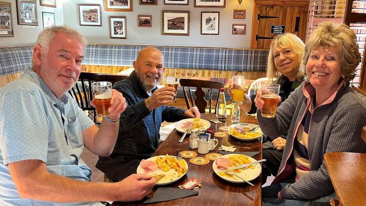 Four customers, two men and two women are sitting at a table in a pub eating ham, egg and chips.
They have paused to raise their glasses of drinks.