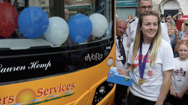 A blonde woman in a white t-shirt wears a medal with a blue ribbon around her neck as she poses next to a stationary bus after a naming ceremony.