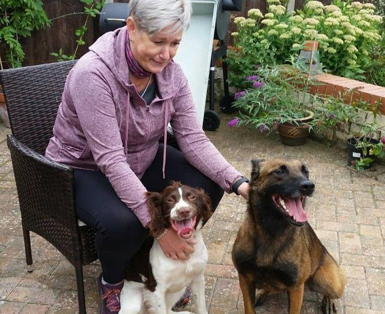 Sharon sits in a chair on the garden patio. She wears a dusty pick hoodie and leans forward with her hands on both dogs. Quinta looks like a alsation cross, and the other dog is some kind of spaniel.
