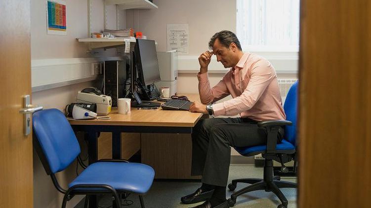 A GP sits at his desk, holding his hand to his head, with an empty patient's chair beside him.