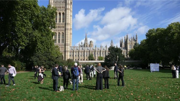 People and dogs on a patch of grass outside the Houses of Parliament in Westminster - a Gothic building. The sky is blue with a few white fluffy clouds.