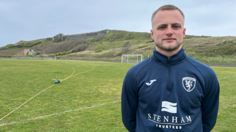 Andrew Mills stands in an Alderney football tracksuit top with the backdrop of a football pitch behind him.