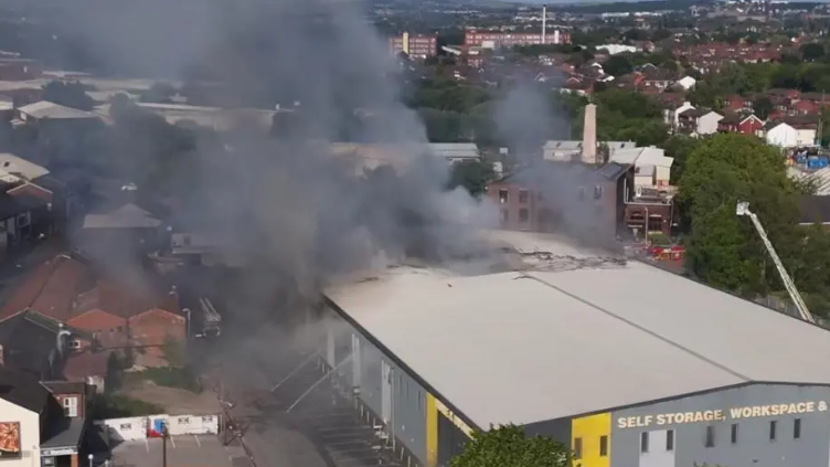 An aerial view of the fire at Storage World, showing the modern, white-rooved building and black smoke pouring from one side
