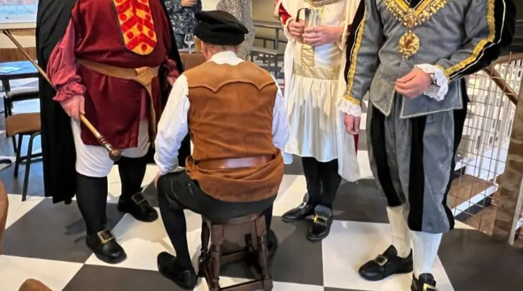 A man in medieval dress sits on a wooden stool with his back to the camera. He is surrounded by three others in medieval dress.