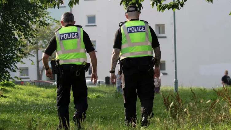Two police officers with their back to the camera. Both are wearing dark clothing and boots with hi-viz jackets over the top with the word police on the back in white writing on a blue background. The officer on the right is wearing a cap. They are walking through long green grass towards a white block of flats with small windows in the side.