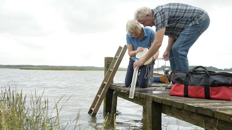Two volunteers standing on a wooden quay sample water on the tidal stretch of the River Alde