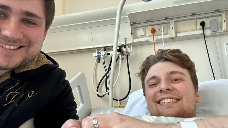 A smiling man in a hospital bed, his hand raised showing a silver ring on his finger, with a smiling man in a black top sitting by his bedside. 