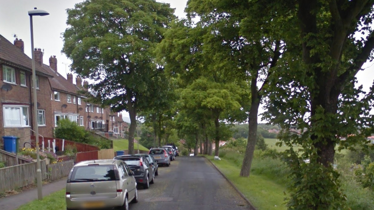 A view straight along Barrowcliff Road in Scarborough, with red-brick houses and cars on the left of the road, and trees and fields to the right.