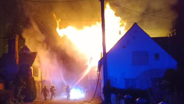 Firefighters spray water from hoses on to a house, which has flames pouring out of it and smoke.