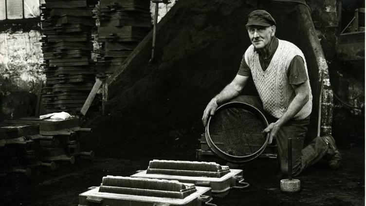 Black and white photo of a man on one knee, holding a round object over a set of foundry moulds. He is looking at the camera and wearing a knitted vest over a short-sleeved shirt and a cloth cap.