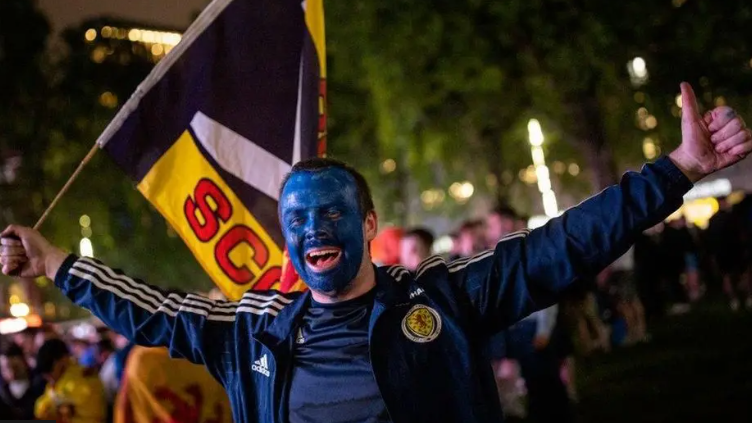 Neach-leantainn sgioba ball-coise na h-Alba a' dèanamh gàirdeachas. Tha aodann air a pheantadh gorm