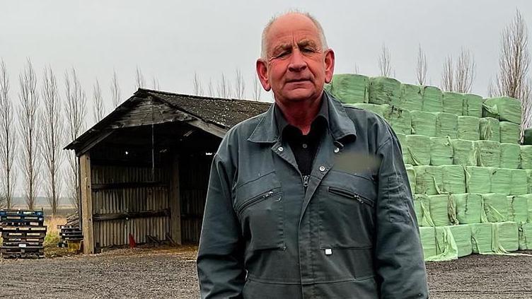 Councillor Julian Kirk wears dark blue overalls. He stands in front of a large barn and bales of animal forage wrapped in green plastic.
