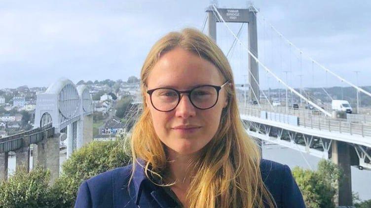 Anna Gelderd is standing outside on a cloudy day with the Tamar and Tamar Bridge behind her. She is smiling slightly and has long blonde hair. She is wearing a navy jacket and a pair of glasses. 